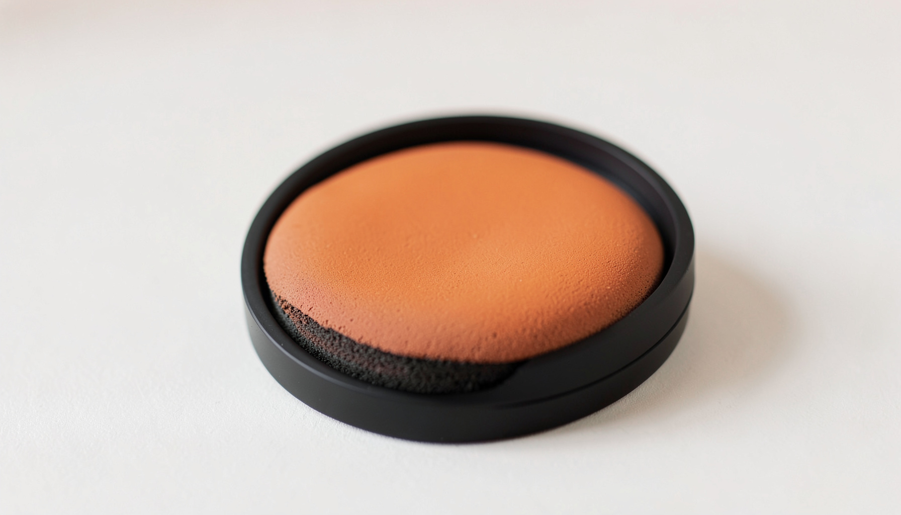 Primer plano de una base de maquillaje en formato cushion compacto negro con esponja, sobre una superficie clara con tonos suaves.
